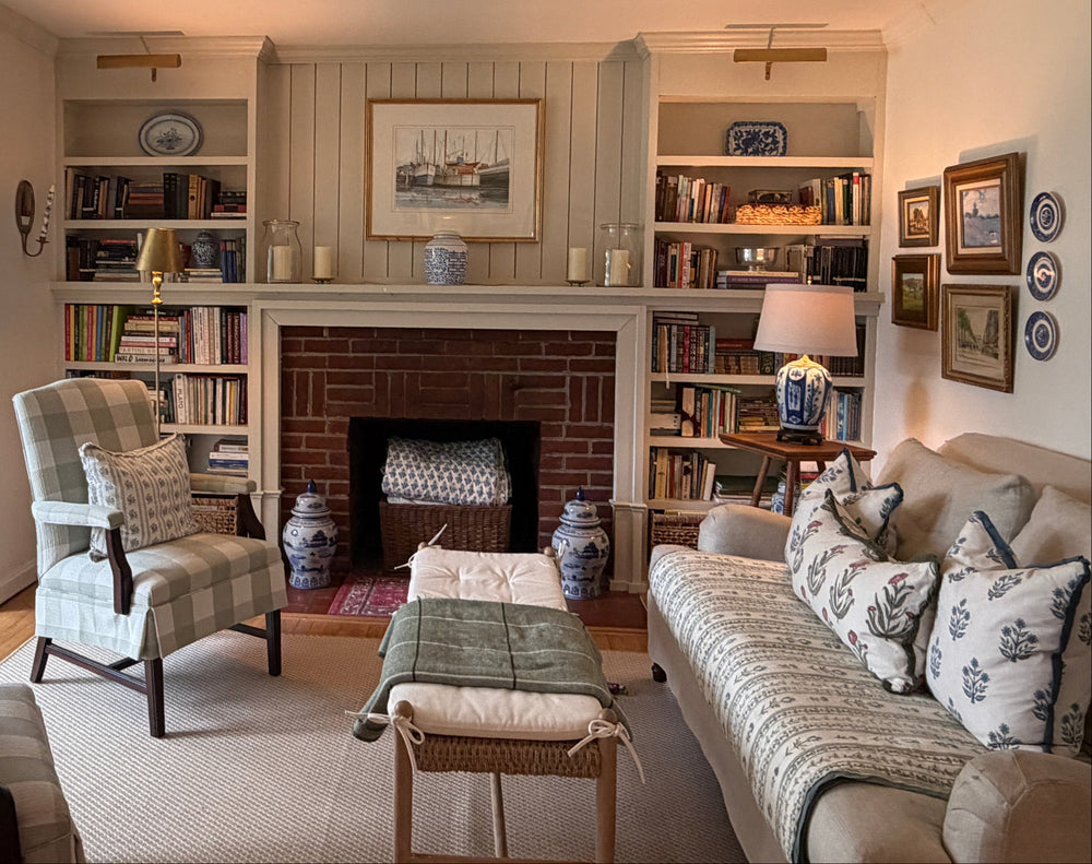 A cozy, traditional living room featuring a central brick fireplace with built-in bookshelves on either side, a beige sofa with patterned pillows, a plaid armchair, and a matching ottoman on a light rug.